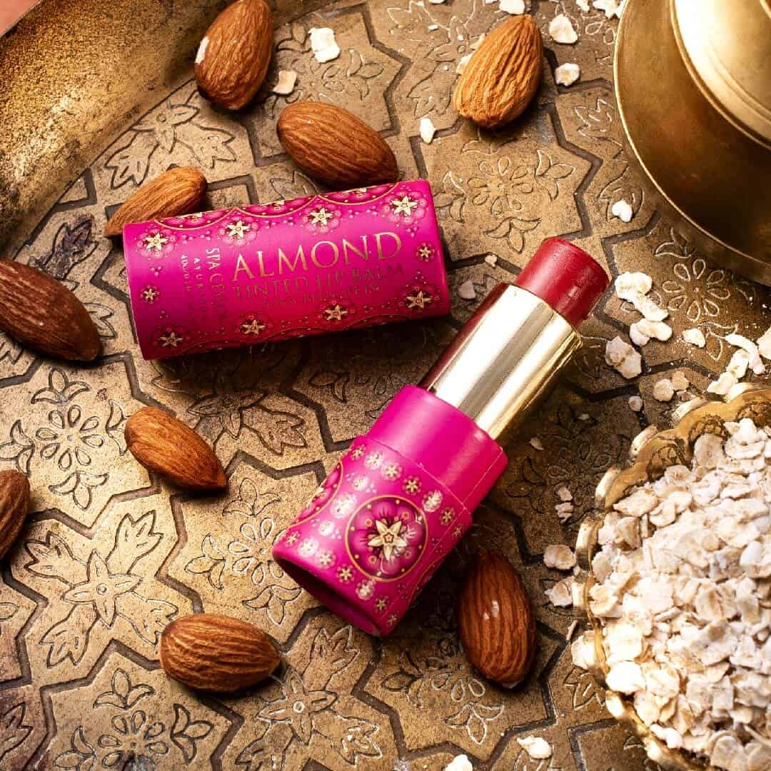 Almond tinted lip balm with floral packaging on decorative tile surface with almonds and gold container.