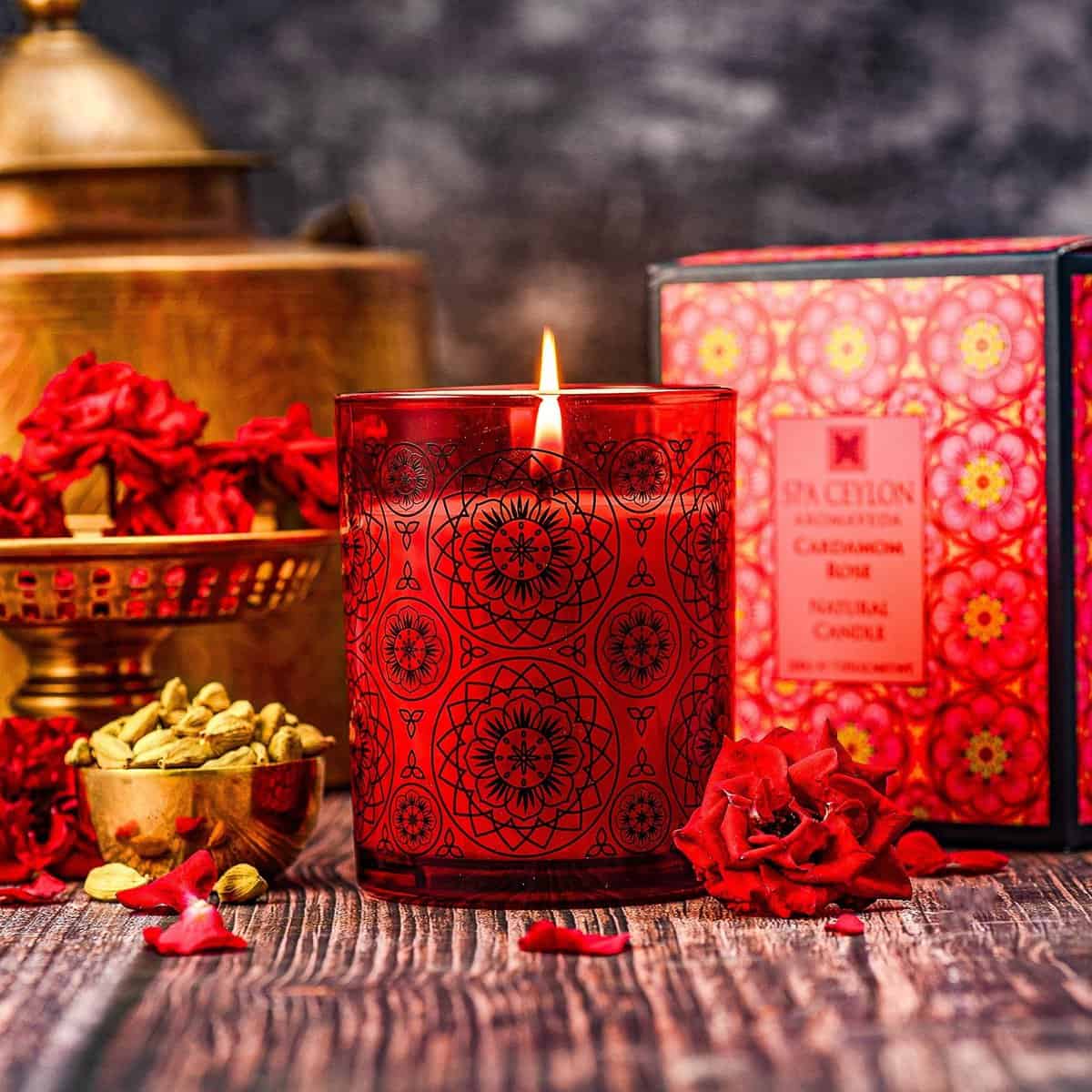 Red scented candle with intricate patterns, surrounded by rose petals, cardamom, and decorative box, creating a relaxing spa ambiance.